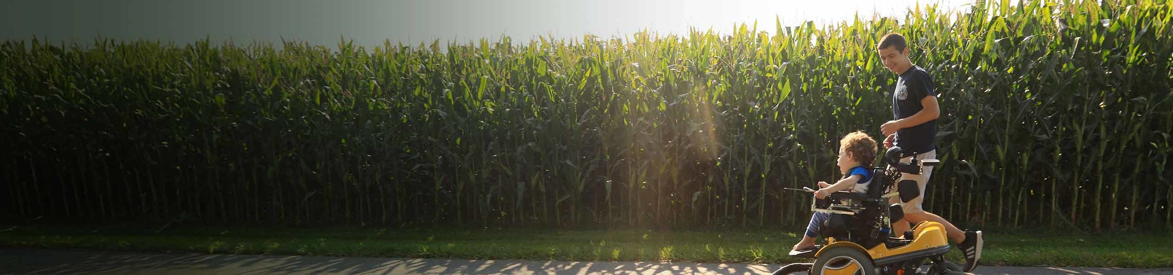 Eli, a child with SMA, in a wheelchair crossing by a cornfield with a male loved one