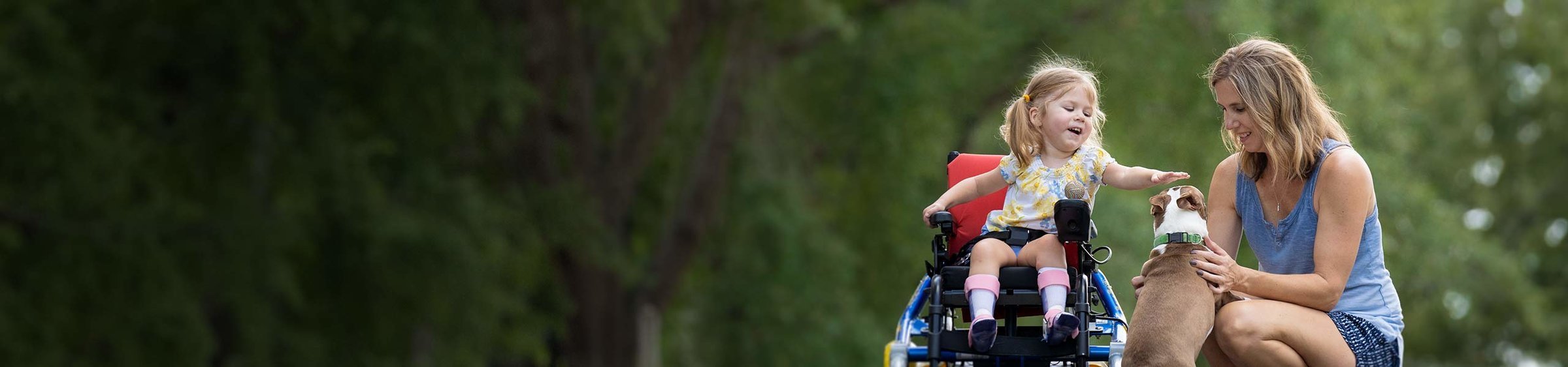 Adalyne, a child with SMA, in wheelchair, eagerly petting a dog as her mother, Patti, holds the dog next to her