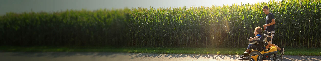 Eli, a child with SMA, in a wheelchair crossing by a cornfield with a male loved one