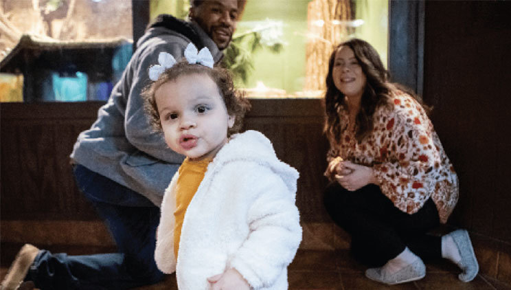 Aniya, a child with SMA, looking forward as her parents, Hailey and Will, gaze at her with pride